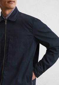 Navy blue zip-up jacket with two front pockets, made of textured fabric. Features a classic collar and subtle stitching details.