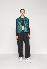 Gilet à imprimé léopard vert avec un col, superposé sur un haut court noir, associé à un pantalon noir et des baskets blanches.