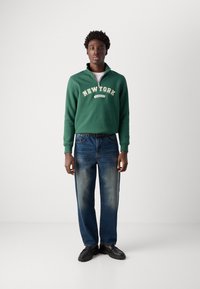 Sweatshirt vert zippé avec "NEW YORK" et "SCHOTT" imprimés en blanc. Associé à un jean en denim bleu clair et des chaussures noires.