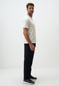 Weißes T-Shirt mit kurzen Ärmeln, kombiniert mit schwarzen Hosen und grauen Turnschuhen. Das Model steht im Profil vor einem schlichten weißen Hintergrund.