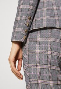 Marynarka w kratę w kolorach szarym, różowym i czarnym. Zbliżenie na zapinany mankiet i starannie skrojony fason. Teksturowana tkanina.