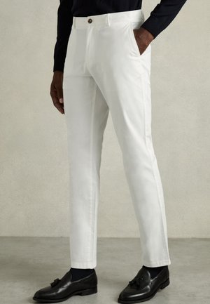 Man draagt slanke witte broek met zijzakken, zwarte top met lange mouwen en zwarte loafers met kwastjes, staand op een grijze vloer.