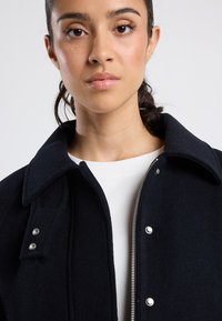 Joven mujer con el cabello oscuro recogido lleva un abrigo de lana negro sobre una camiseta blanca de cuello redondo, contra un fondo claro y liso.