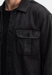 Zwarte denim shirt met een knoopsluiting, een borstzak met klep en een zachte textuur. De kraag en de stiknaad zijn zichtbaar.