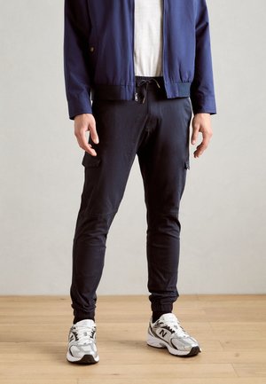 Mann trägt marineblaue Jogginghosen, weiße Sneaker, weißes T-Shirt und eine marineblaue Zip-Up-Jacke und steht auf einem Holzboden.