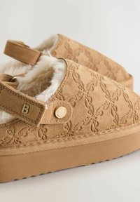 Hellbeige Wildleder-Clogs mit eingeprägtem Blumenmuster, weichem Innenfutter, verstellbarem Riemen und Gummisohle mit strukturiertem Grip.