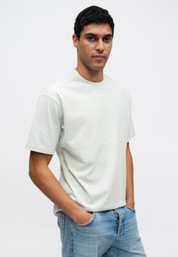 Ανοιχτό γκρι κοντομάνικη μπλούζα με στρογγυλή λαιμόκοψη. Κατασκευασμένη από απαλό ύφασμα, με διακριτικό λογότυπο στο γιακά. Φορεμένη με μπλε τζιν παντελόνι.