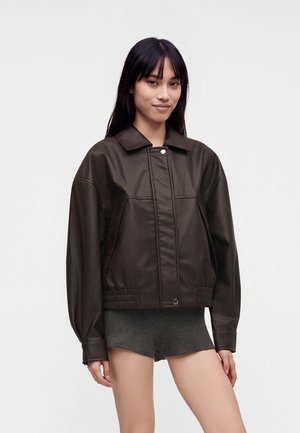 Mujer con una chaqueta de cuero marrón oscuro y pantalones cortos grises, de pie frente a un fondo claro y liso, mirando hacia adelante con una ligera sonrisa.