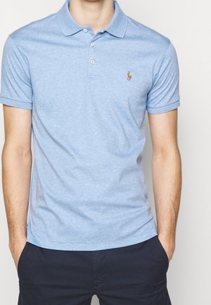 Mann trägt ein hellblaues kurzärmeliges Poloshirt mit zwei Knöpfen und kleinem Pferdelogo auf der Brust, kombiniert mit dunkler Hose.