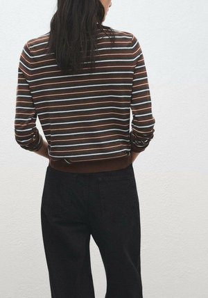 Woman with long dark hair wearing a brown, white, and blue striped long-sleeve top and black pants, standing against a white wall.