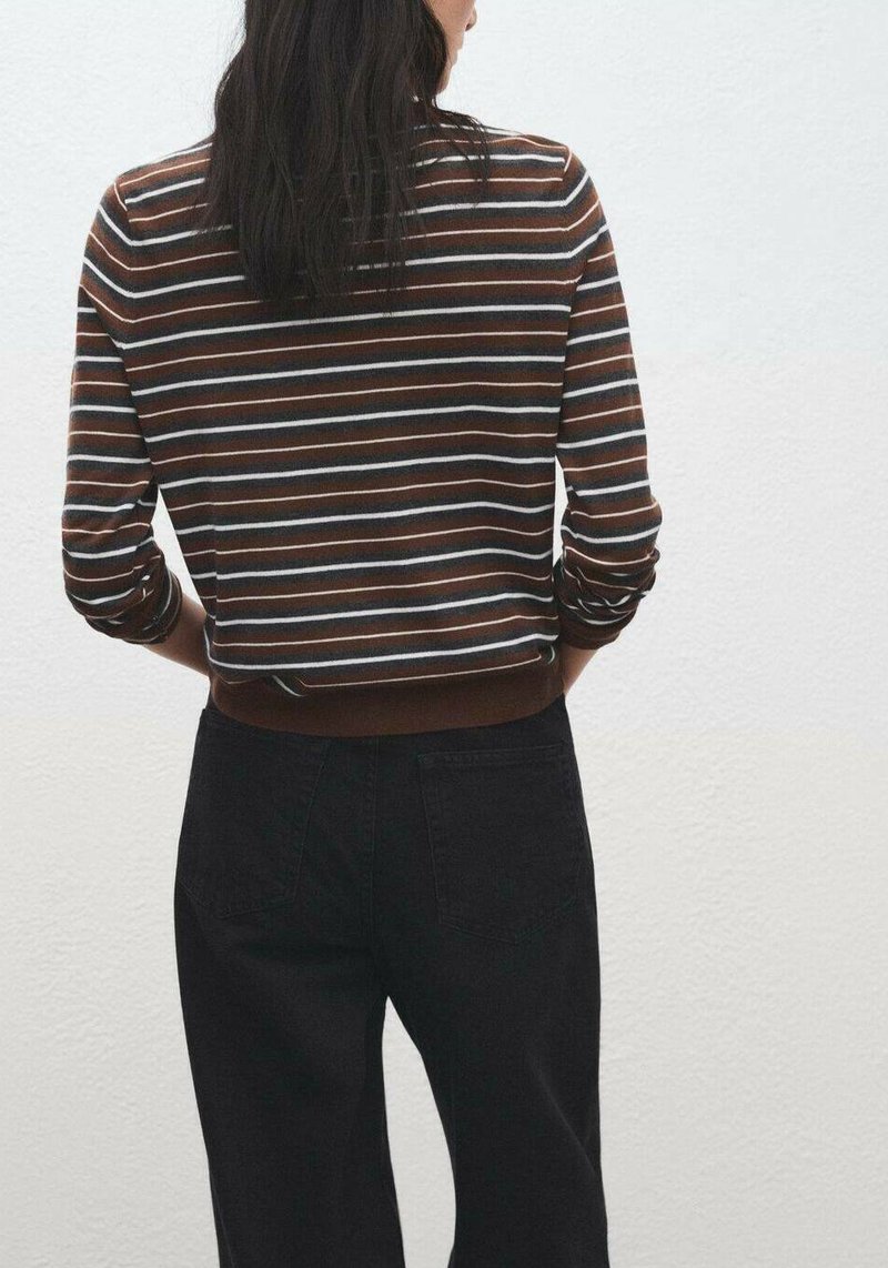 Femeie cu păr lung și închis la culoare, purtând un top cu mâneci lungi cu dungi maro, alb și albastru și pantaloni negri, stând în picioare lângă un perete alb.