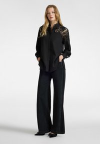 Camicia nera con bottoni e dettagli in pizzo sulle spalle e sulle maniche, abbinata a jeans neri a vita alta e gamba larga e scarpe nere a punta.