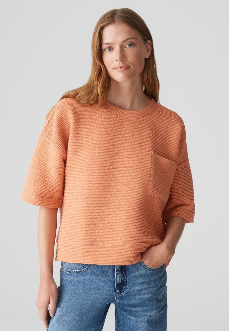 Maglione a maniche corte dallo stile rilassato in tessuto peach testurizzato; presenta un collo rotondo e una tasca frontale discreta; abbinato a jeans blu.