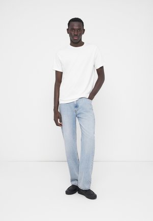 Camiseta blanca de manga corta, vaqueros rectos azul claro, zapatos negros sin cordones, sobre un fondo neutro. Conjunto informal con detalles mínimos.