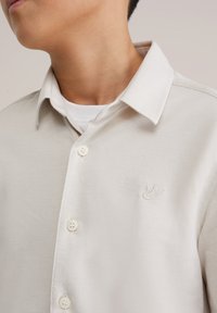 Világos bézs gombos ing, texturált anyagból készült, klasszikus gallérral és egy kis hímzett logóval a mellrészen.