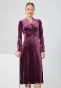 Egy hosszú ujjú, mélylila velúr midi ruhát viselő nő, V-kivágással, aki egyik kezét egy fehér felületen nyugtatja.