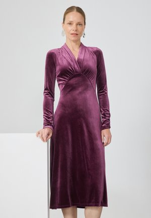 Kvinde iført en langærmet, dyb lilla velour midi-kjole med V-hals, stående med den ene hånd hvilende på en hvid overflade.