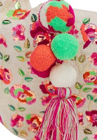 Geweven decoratief stuk met meerkleurige pompons (groen, wit, oranje) en een roze en witte tassel, op een beige bloemrijke stof.