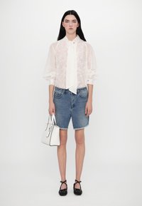 Blusa bianca trasparente con dettagli a fiocco, abbinata a pantaloni shorts di jeans blu. La modella tiene una borsa bianca e indossa delle ballerine nere con borchie.