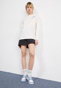 Light cream long-sleeve shirt with blue logo, paired with a black skirt. White and light blue sneakers, green-striped socks. Simple, casual look.