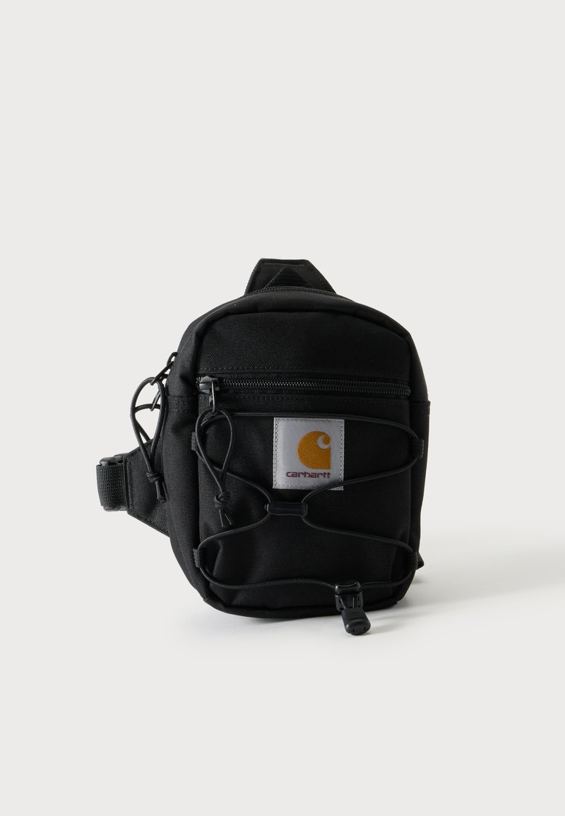 Συμπαγής μαύρη τσάντα μέσης Carhartt με εμπρόσθια τσέπη με φερμουάρ, ρυθμιζόμενο λουρί και ελαστική λεπτομέρεια με κορδόνι σε μονόχρωμο φόντο.