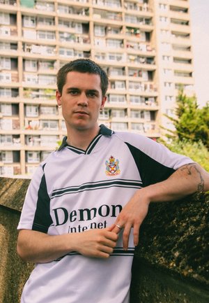 Witte en zwarte T-shirt met korte mouwen en een logo, voorzien van horizontale strepen en teksten. Model leunt tegen een ruwe betonnen muur.