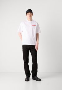 Hvit bomulls t-skjorte med rød "CALVIN KLEIN" tekst på brystet, parret med svarte jeans og svarte sko, toppet med en svart caps.