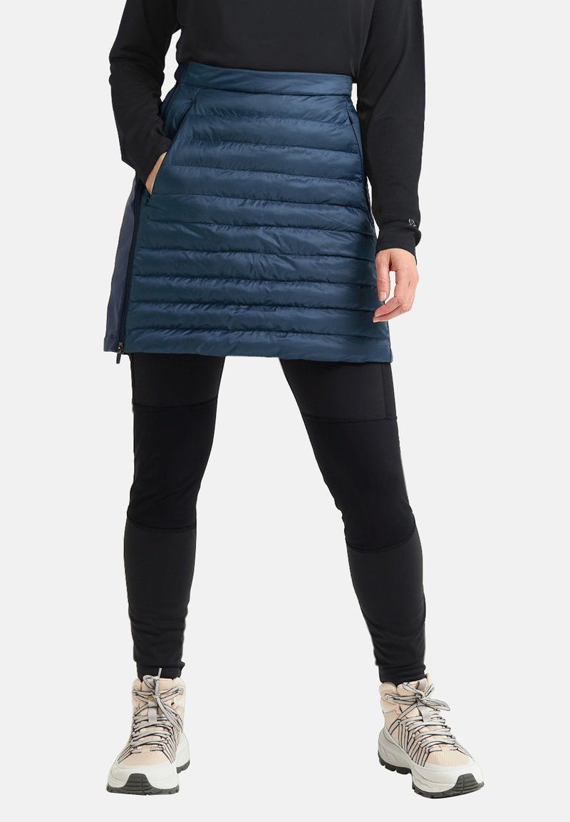 Marineblauwe pufferrok met horizontale stiksels, voorzien van zijzakken en een rits. Gecombineerd met zwarte leggings en lichtgekleurde wandelschoenen.