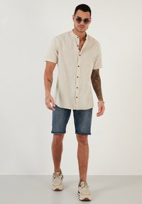 Cămașă scurtă de culoare bej deschis, cu guler bandă și închidere cu nasturi, purtată cu pantaloni scurți din denim și adidași bej. Textură netedă, stil casual.