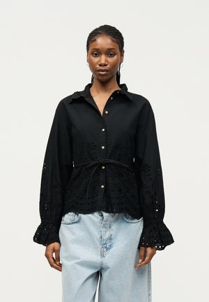 Žena s vrkočmi, oblečená v čiernej blúzke na gombíky s očkovými detailmi a svetlomodrých vysokoťahavých džínsoch, stojaca pred jednoduchým pozadím.