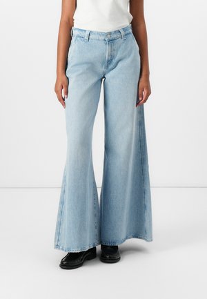 Jean large bleu clair en denim, avec taille haute, fermeture à bouton et décoloration subtile. Une paire de chaussures noires vient parfaire le look.