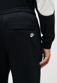 Κοντινό πλάνο σε μαύρο παντελόνι φόρμας Nike με πίσω τσέπη και λευκό κεντημένο λογότυπο, φορεμένο από άτομο με μαύρο τοπ και λευκή μπαλώνα στον αγκώνα.