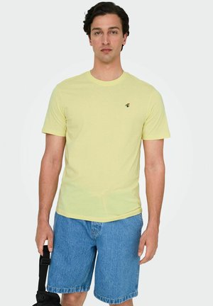 Gelbes Baumwoll-T-Shirt mit Rundhalsausschnitt, kurzen Ärmeln und einem kleinen bestickten Pflanzenlogo auf der Brust, kombiniert mit blauen Jeansshorts.