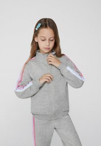 Šedá mikina na zip s vysokým límcem a růžovými a modrými pruhy na rukávech. Měkká textura, přiléhavý střih, spárovaná s odpovídajícími kalhotami.