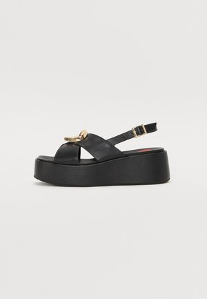 Black leather platform sandals with a cross-strap design and gold-tone hardware detail. Features an adjustable ankle strap and textured sole.