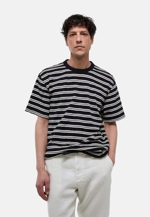 Homme aux cheveux courts et bouclés, portant une chemise à manches courtes rayée noir et blanc et un pantalon blanc, debout avec une main dans la poche.
