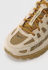 Μπεζ αθλητικό παπούτσι με υλικά σουέτ και δίχτυ, με κίτρινους και καφέ κορδόνια, ανάγλυφη σόλα και εμφανές λογότυπο της Nike.