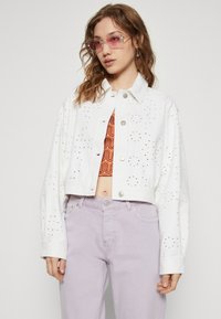 Chaqueta de mezclilla blanca recortada con detalles perforados, botones plateados y un ajuste relajado, combinada con jeans lavanda y una blusa naranja con estampado.
