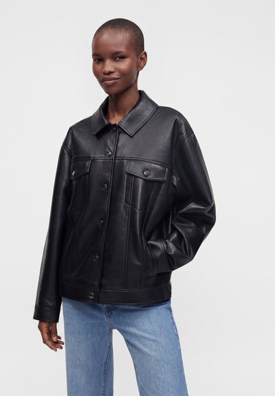 Personne portant une veste noire boutonnée en similicuir avec des poches devant et un jean bleu clair, debout devant un fond uni et clair.