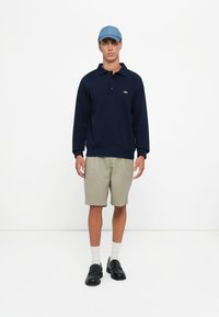 Marineblauer Pullover mit grünem Logo, beigen Shorts, weißen Socken und schwarzen Slippers, abgerundet mit einer hellblauen Mütze.