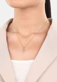 Złota naszyjnik z krzyżykiem zawieszką, ozdobioną małymi okrągłymi białymi kamieniami, noszony przez osobę w beżowej marynarce i białej bluzce.