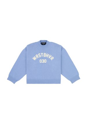 Femme portant un pull oversize bleu clair avec un texte, une jupe mini crème, des chaussures beiges à petits talons, des lunettes et des boucles d’oreilles créoles, debout les mains jointes.