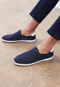 Mörkblå canvas slip-on-skor med vit gummisula, med en kontrasterande vit häl och en liten tri-färgad logotyp nära fotvalvet.