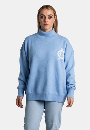 Junge Frau mit glattem, blondem Haar, die einen hellblauen Rollkragenpullover und hellblaue Jeans trägt, vor einem schlichten Hintergrund stehend.