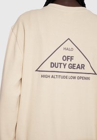 Beige texturerad sweatshirt med en bakre grafik som visar en triangulär kontur och svart text: "HALO OFF DUTY GEAR HIGH ALTITUDE LOW OPENING."