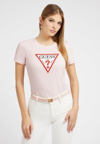 Rožinė trumpa rankovė marškinė su trikampiu raudonu logotipu, ant kurio yra žodis "GUESS" ir klausimo ženklas, derinami su baltais džinsais ir rožiniu diržu.
