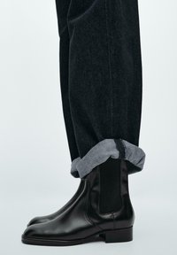 Stivaletti Chelsea in pelle nera con pannelli elastici laterali, abbinati a jeans scuri risvoltati e un bordo in denim grigio, che mostrano un design elegante.