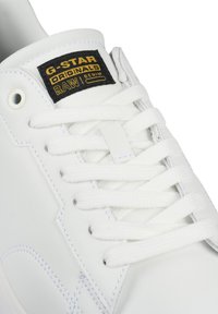 Vita lädersneakers med platta vita snören, med en svart etikett med guldtext. Slät yta och sömnadsdetaljer synliga.