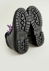 Bottes ankle en suede noir avec des semelles en caoutchouc texturé à forte adhérence. Les lacets violets soulignent le design. Nom de la marque "NEXT" sur les semelles.