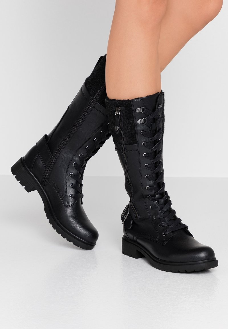 Bottes noires à lacets mi-mollet avec des zips latéraux, des boucles et des poignets texturés, portées sur des jambes croisées devant un fond uni.